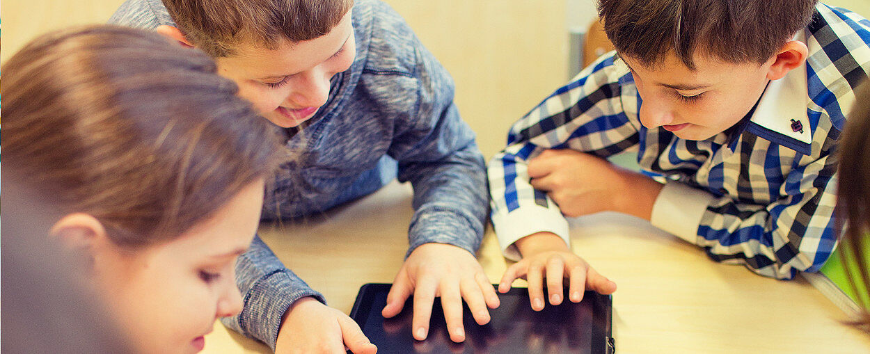 junge Schülerinnen und Schüler, fröhlich und konzentriert, bei gemeinsamer Gruppenarbeit mit iPad an einem Tisch sitzend