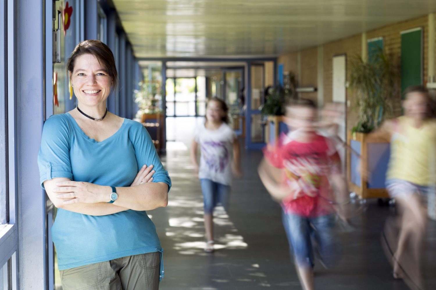 Portrait einer lächelnden Grundschullehrerin, an einer Flurwand lehnend, laufende Kinder im Hintergrund